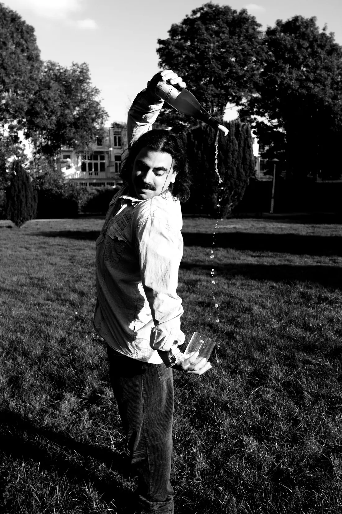 Aitor pouring cider from above his head into a glass held at his hip, in a park