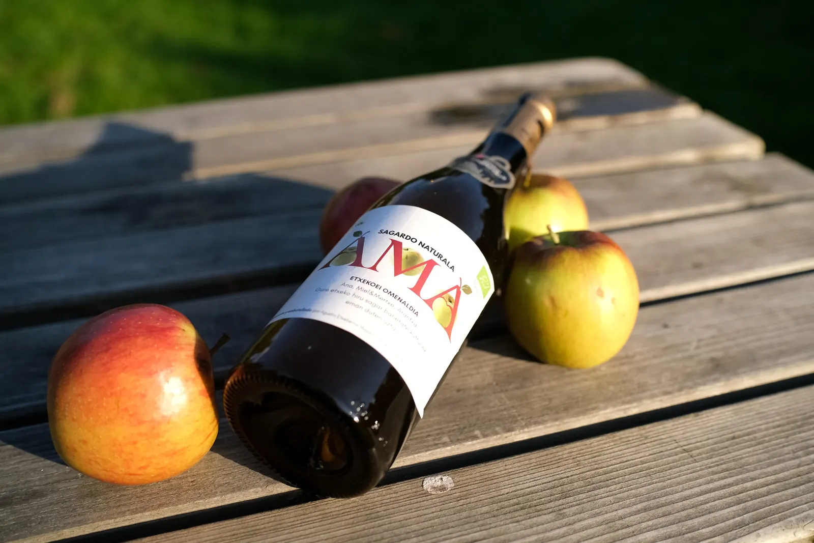 AMÁ natural cider bottle lying on a wooden table between red and yellow apples